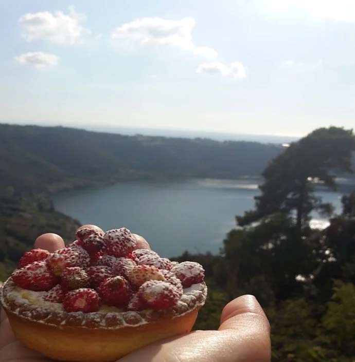 foto di una mano che tiene un dolce con guarnizione di fragoline di Nemi con sfondo il lago